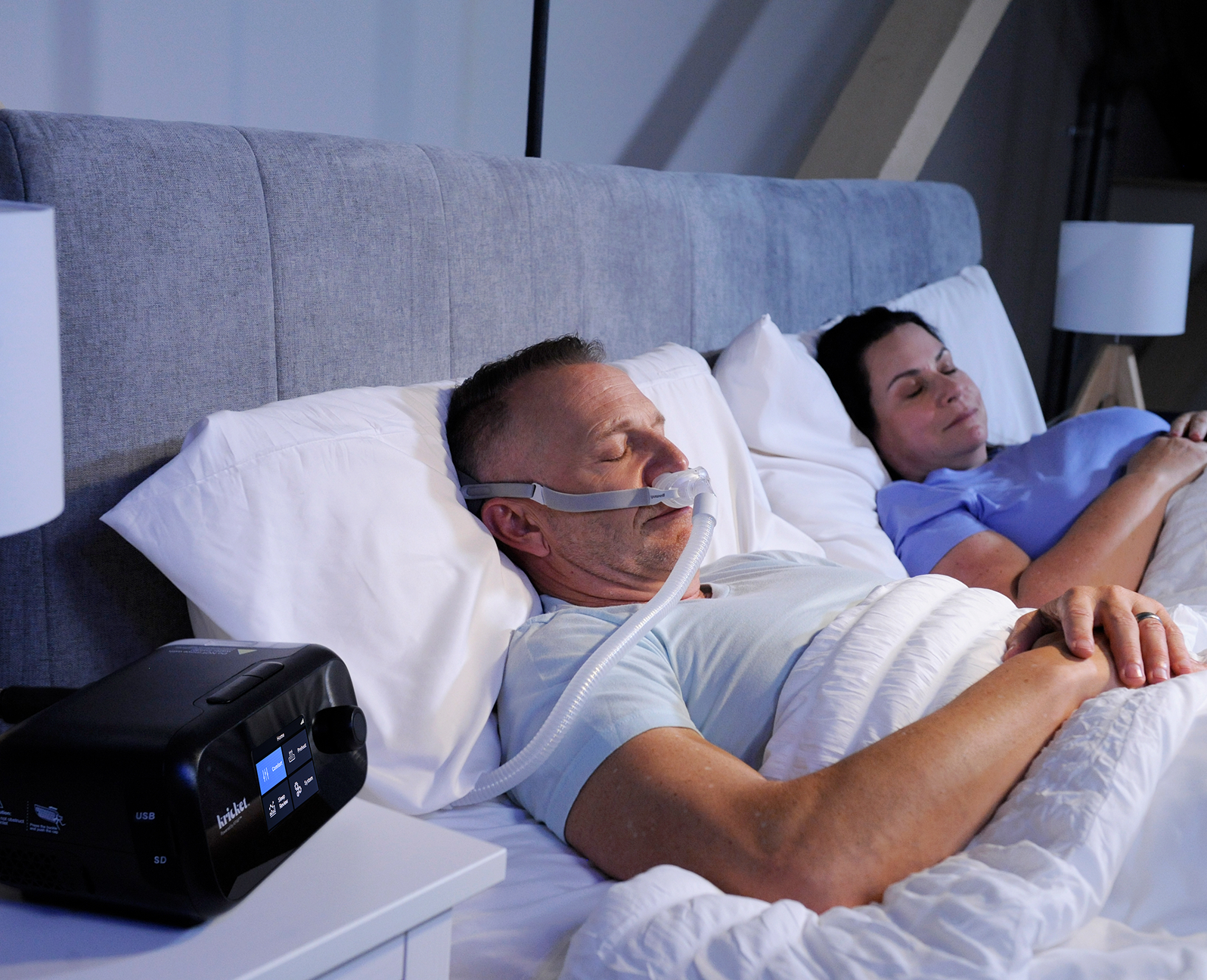 Man sleeping with his Kricket PAP device on the nightstand while his partner sleeps beside him