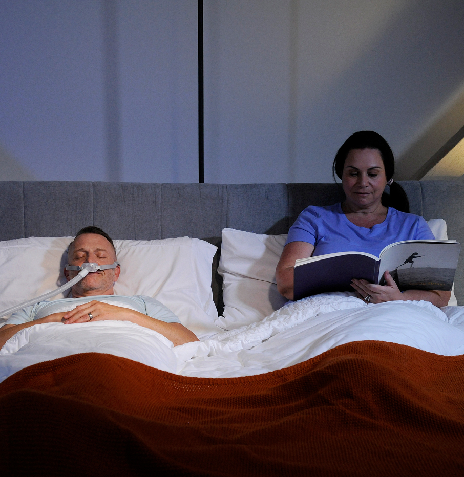 Man sleeping with his PAP mask on while his partner reads beside him undisturbed